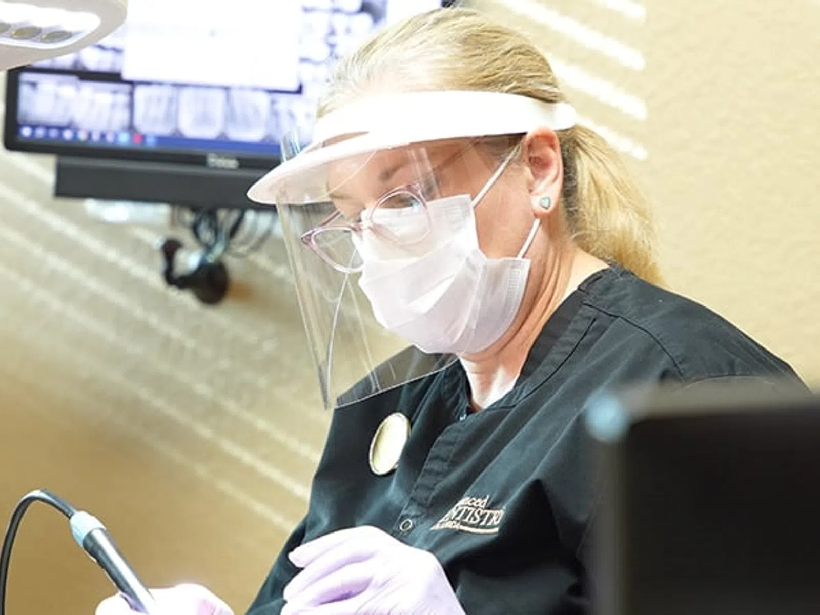 Dentist wearing face shield and mask performing dental procedure in a clinic with digital screen in background
