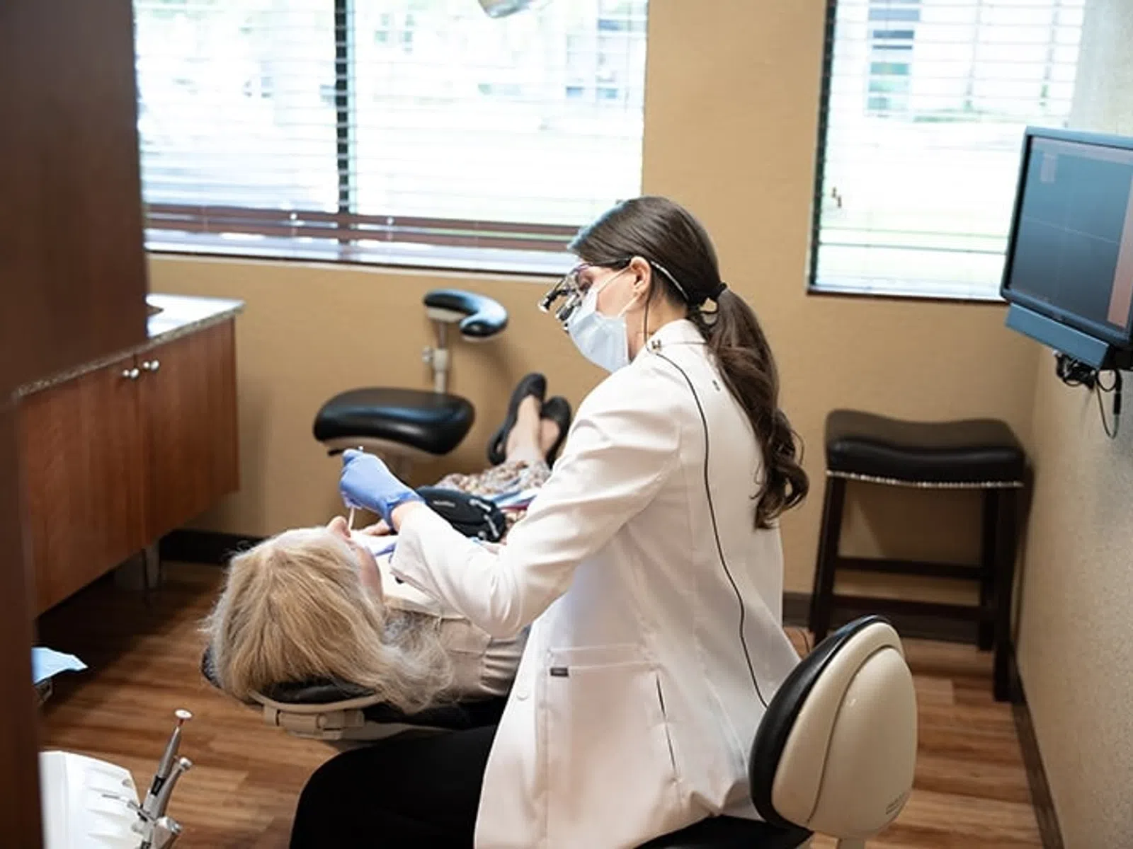 Dentist wearing a mask and gloves treating a patient in a modern dental clinic with equipment and natural light.