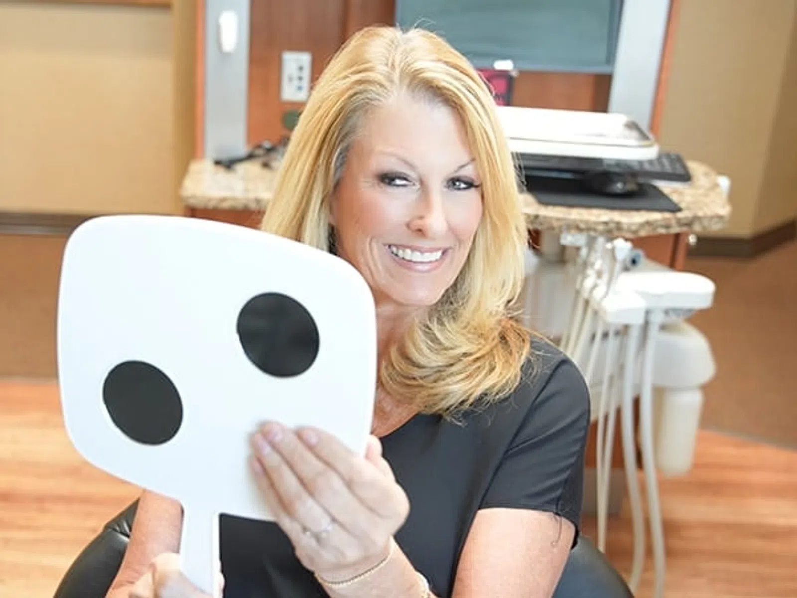 Smiling woman with blonde hair holding a dental mirror with black circles in a dental office setting.