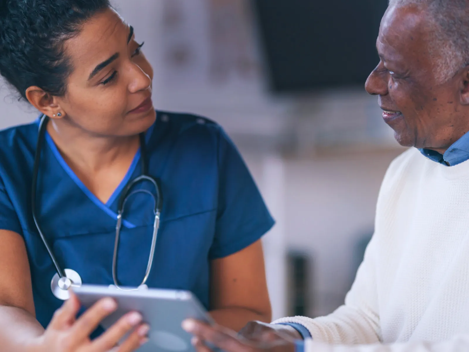 Female nurse in blue scrubs consulting with elderly male patient using a tablet in a medical setting