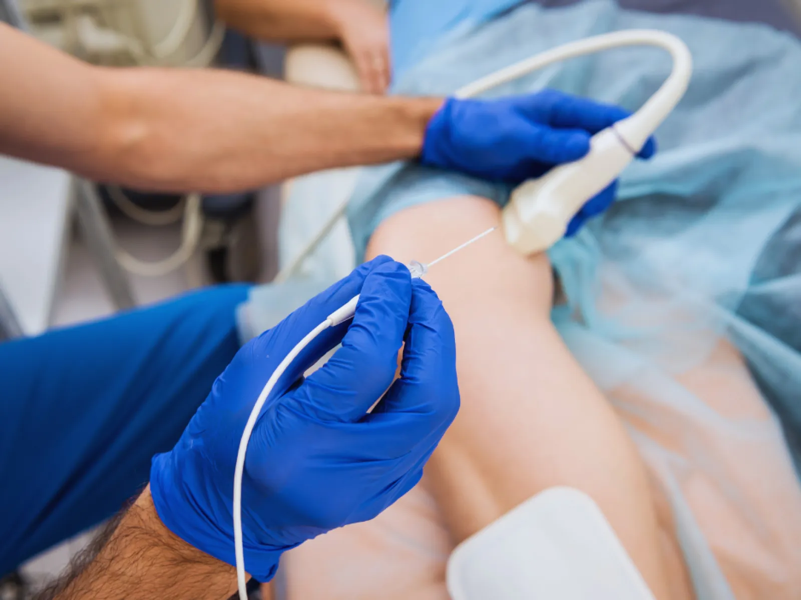 Medical professional performing ultrasound-guided injection on patient's knee wearing blue gloves in clinic.