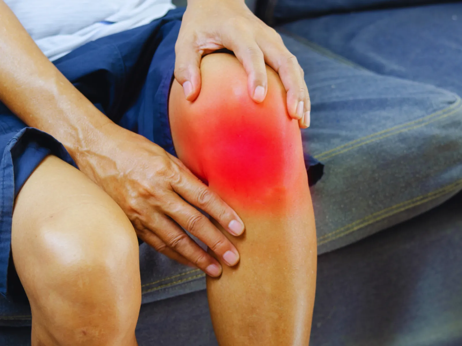 Person holding their knee with visible red area indicating pain or inflammation while sitting on couch.