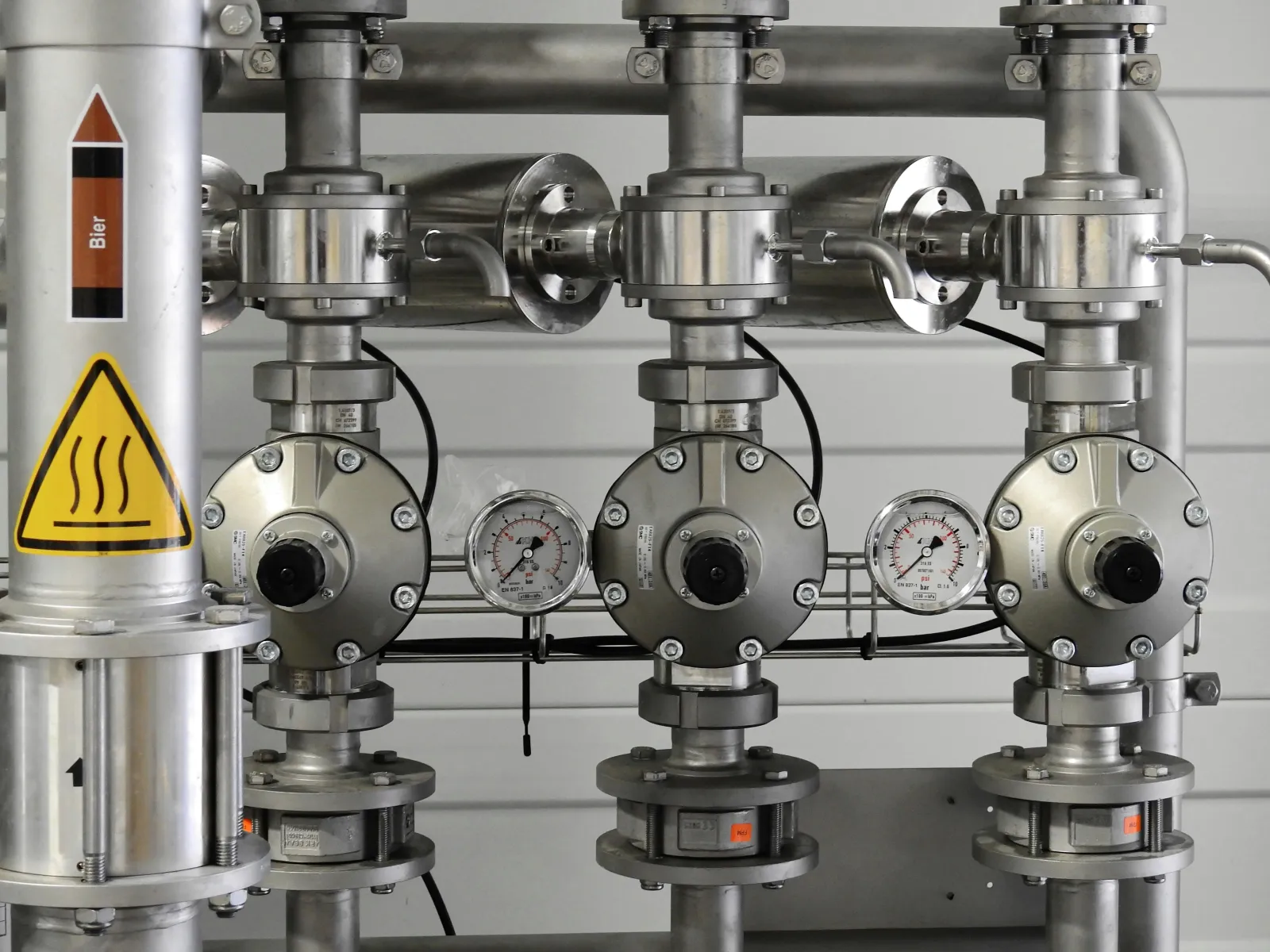 Close-up of industrial stainless steel pipe system with pressure gauges and hot liquid warning sign.