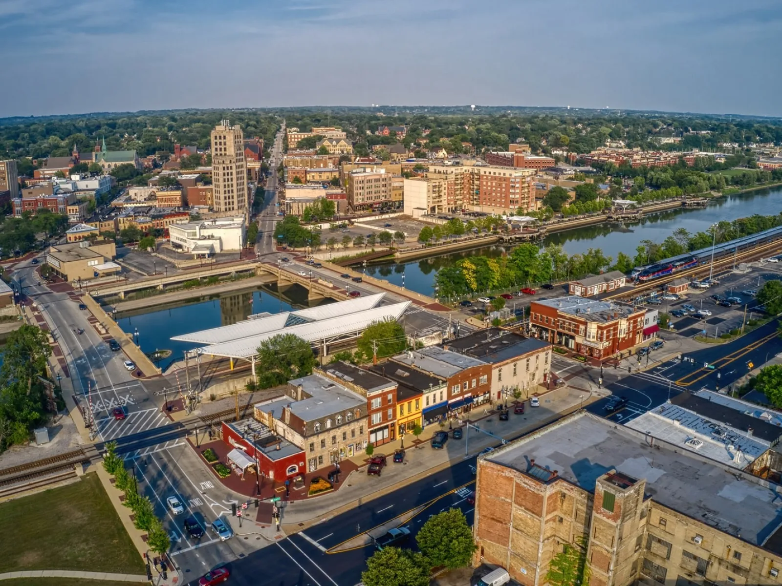 Aerial view of a riverfront city with bridges, historic buildings, train tracks, and green trees under a blue sky.