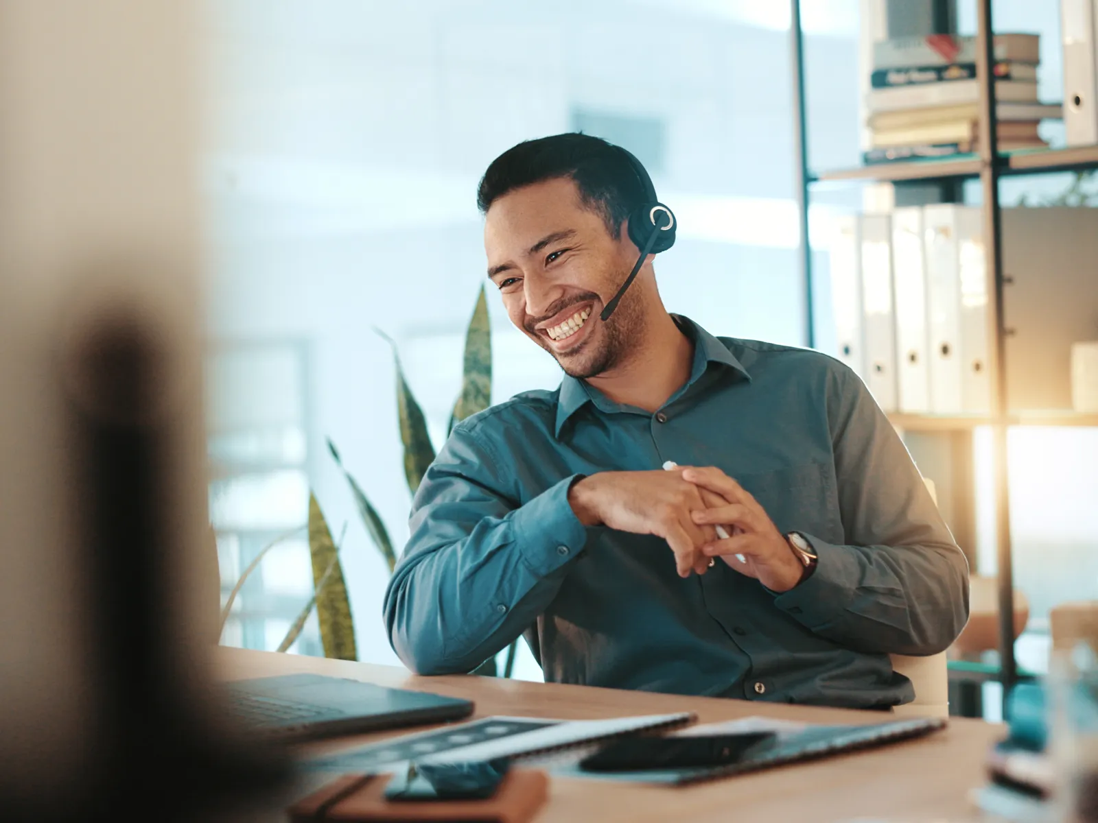 Smiling customer support representative wearing headset working at desk with laptop in modern office.