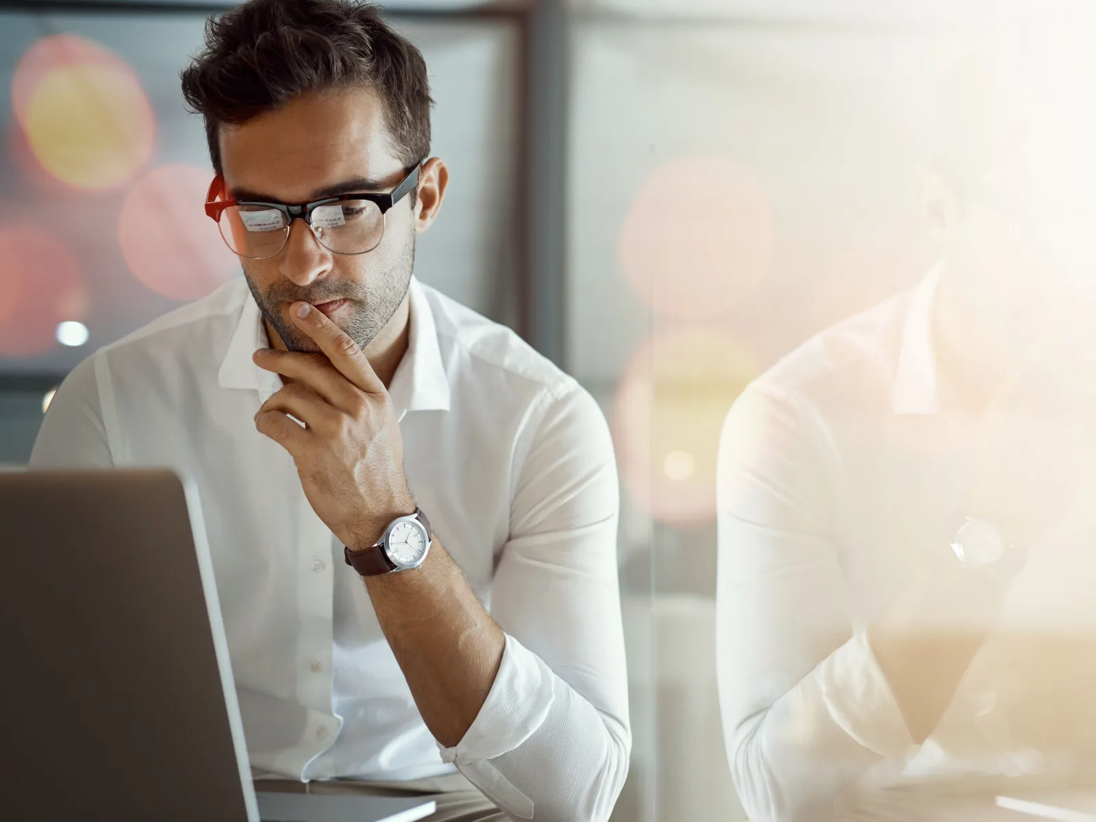 Young man in glasses working focused on laptop in modern office with reflection and warm lighting.