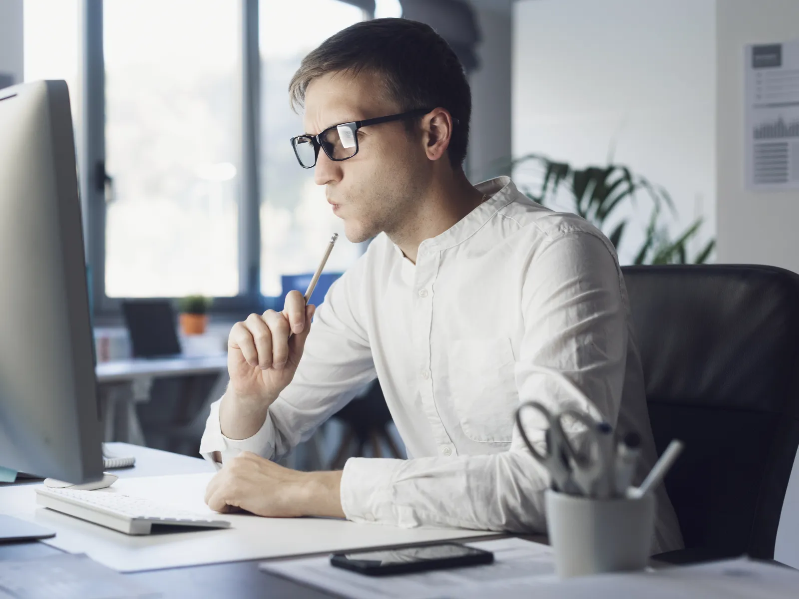 Man in white shirt and glasses thoughtfully working on computer in bright office environment.