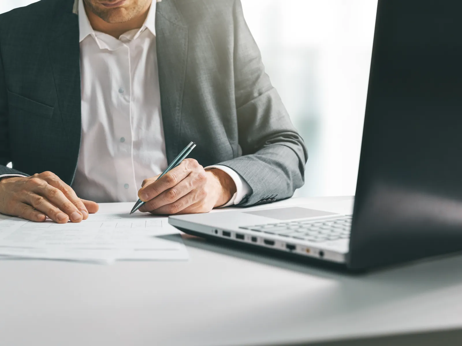 Businessman in suit signing documents at desk with laptop in a modern office environment.