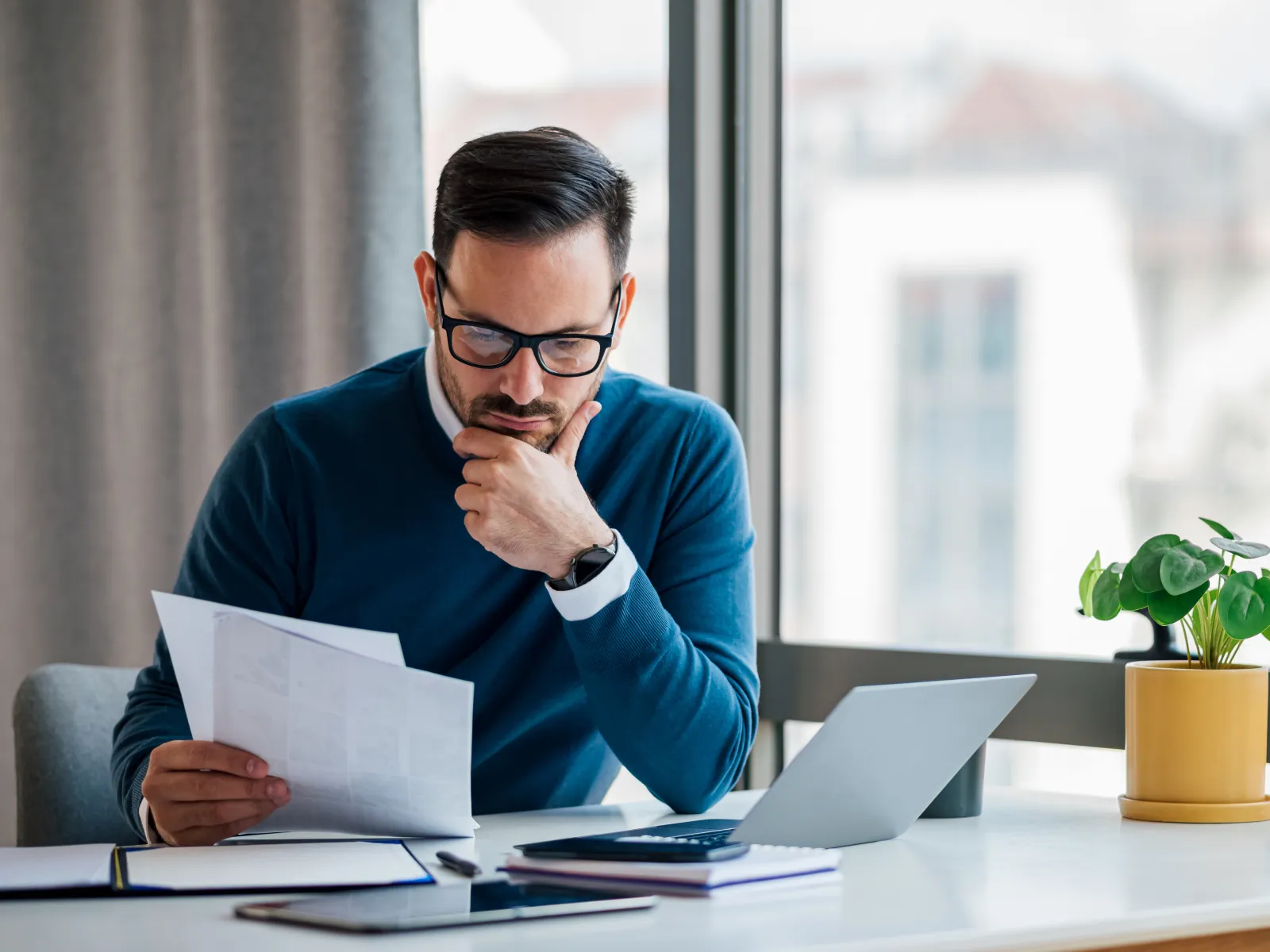 Man in glasses studying documents at a desk with a laptop and potted plant by the window in a bright office.