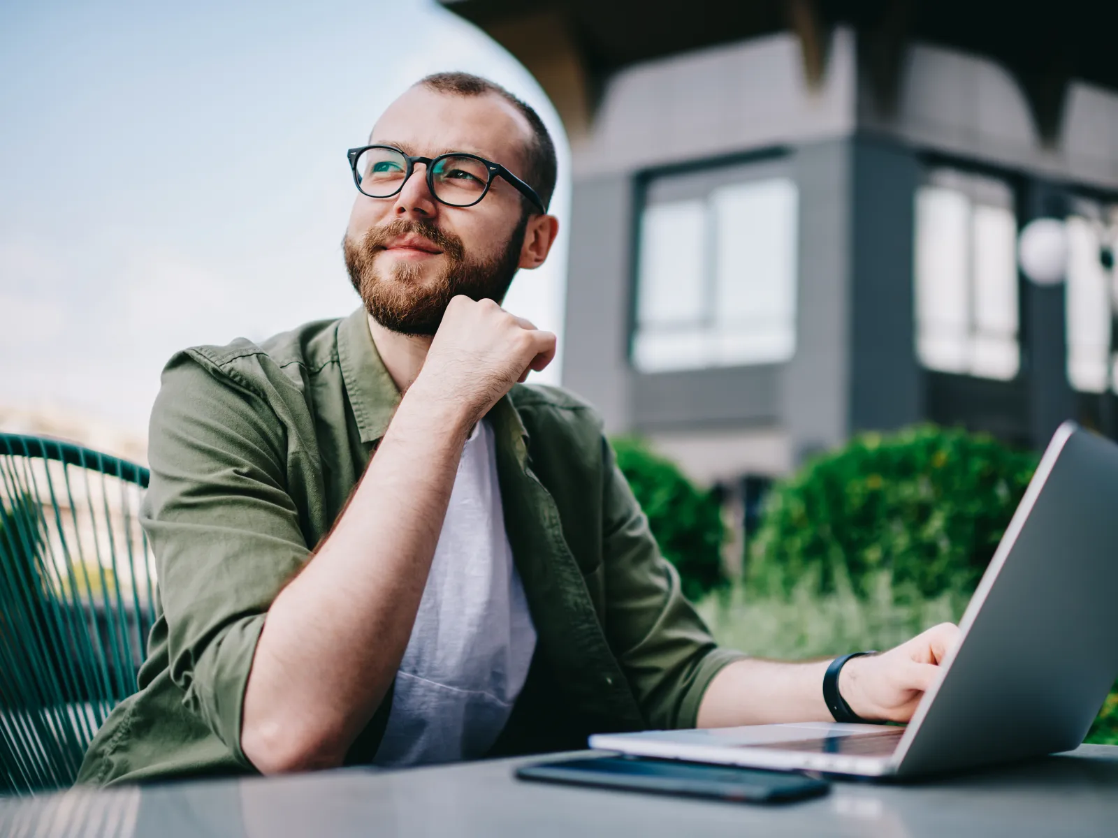 Young man with glasses working on laptop outdoors looking thoughtfully into the distance on a sunny day