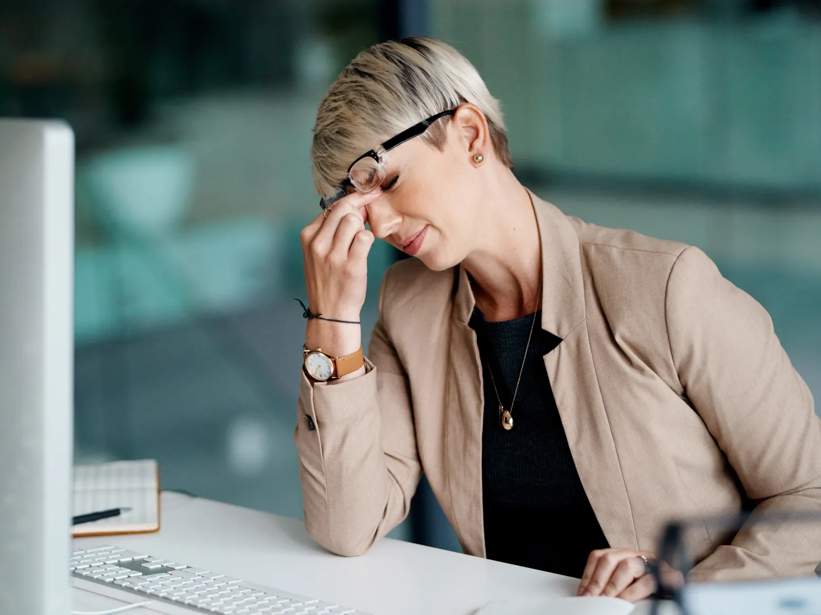 Frustrated woman in business attire rubbing her forehead while sitting at a desk with computer keyboard.