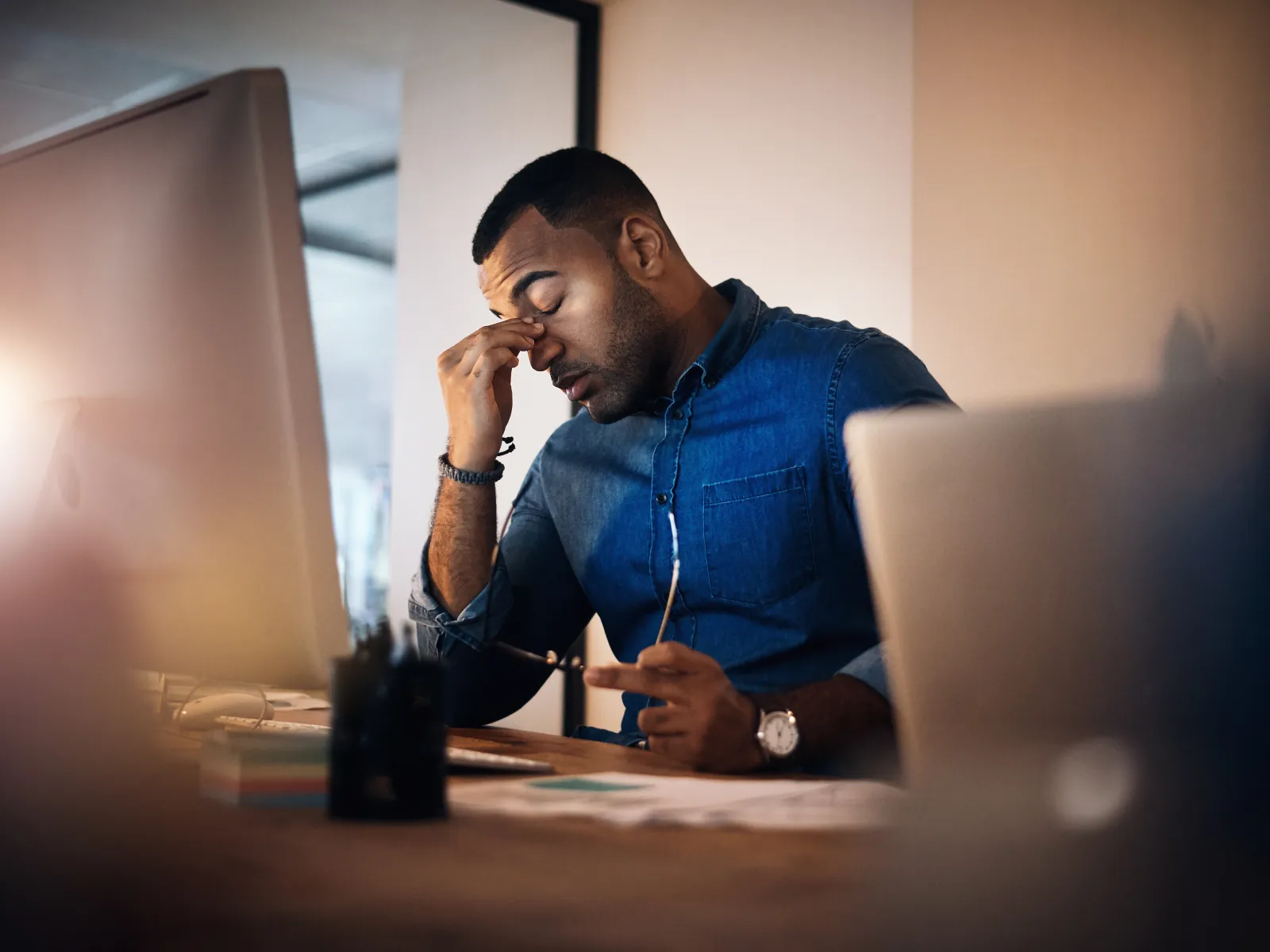 Man in blue shirt working at desk rubbing eyes looking stressed or tired with computer and papers around.