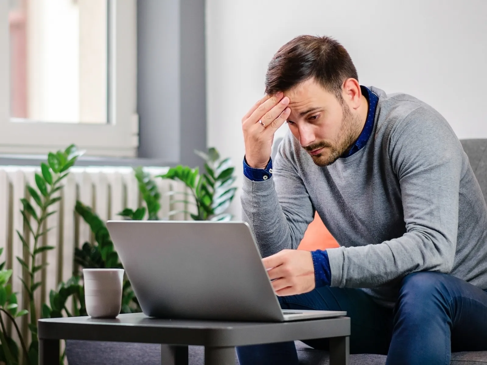 Man in gray sweater looking stressed while working on laptop at home with cup and plants nearby.
