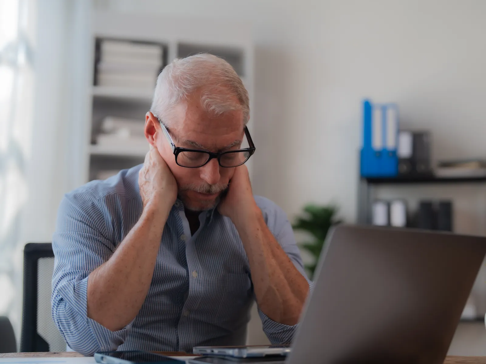 Mature man wearing glasses massaging neck while working on laptop at desk in home office.
