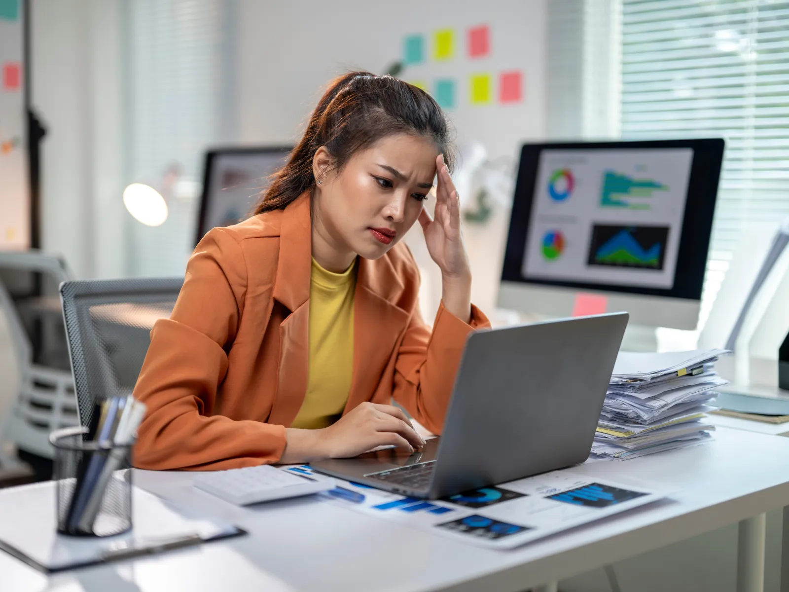 Stressed businesswoman in orange blazer working on laptop in office surrounded by papers and charts.