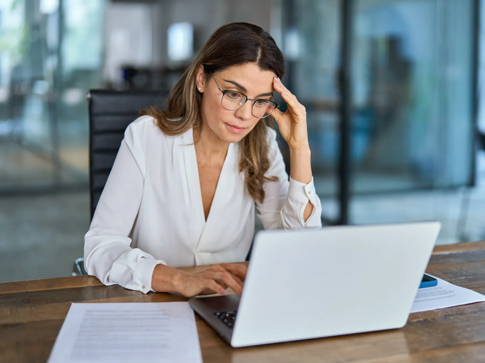 Professional woman in glasses working on laptop with documents at wooden desk in modern office environment