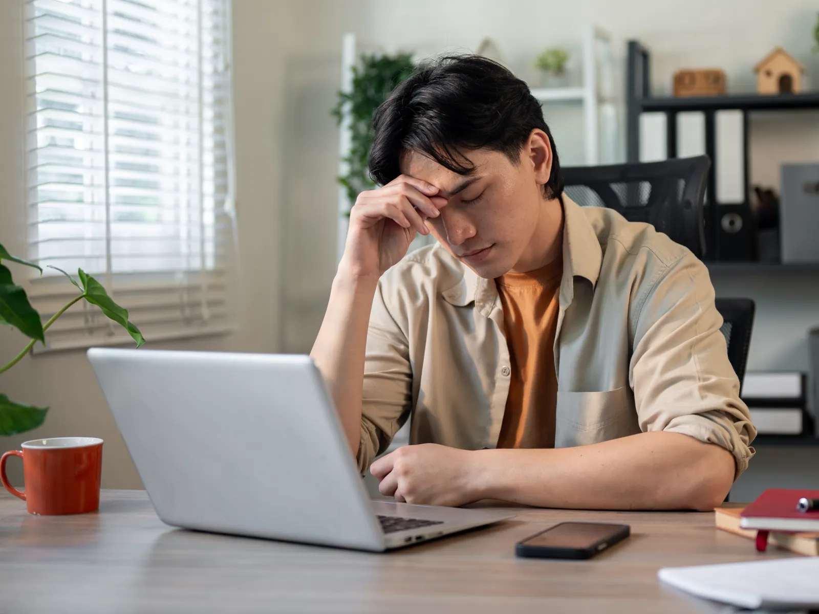 Young man looking stressed while working on a laptop at a desk with notebooks and smartphone.