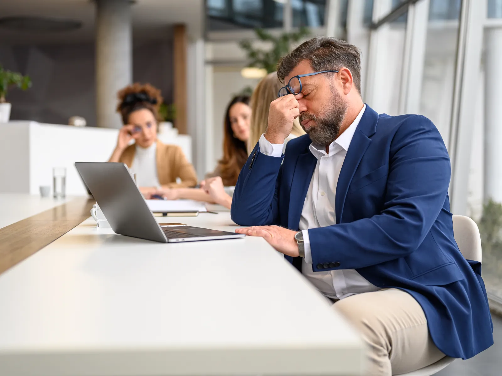 Stressed businessman in blue suit sitting at desk with laptop, rubbing eyes, with two women blurred in background.