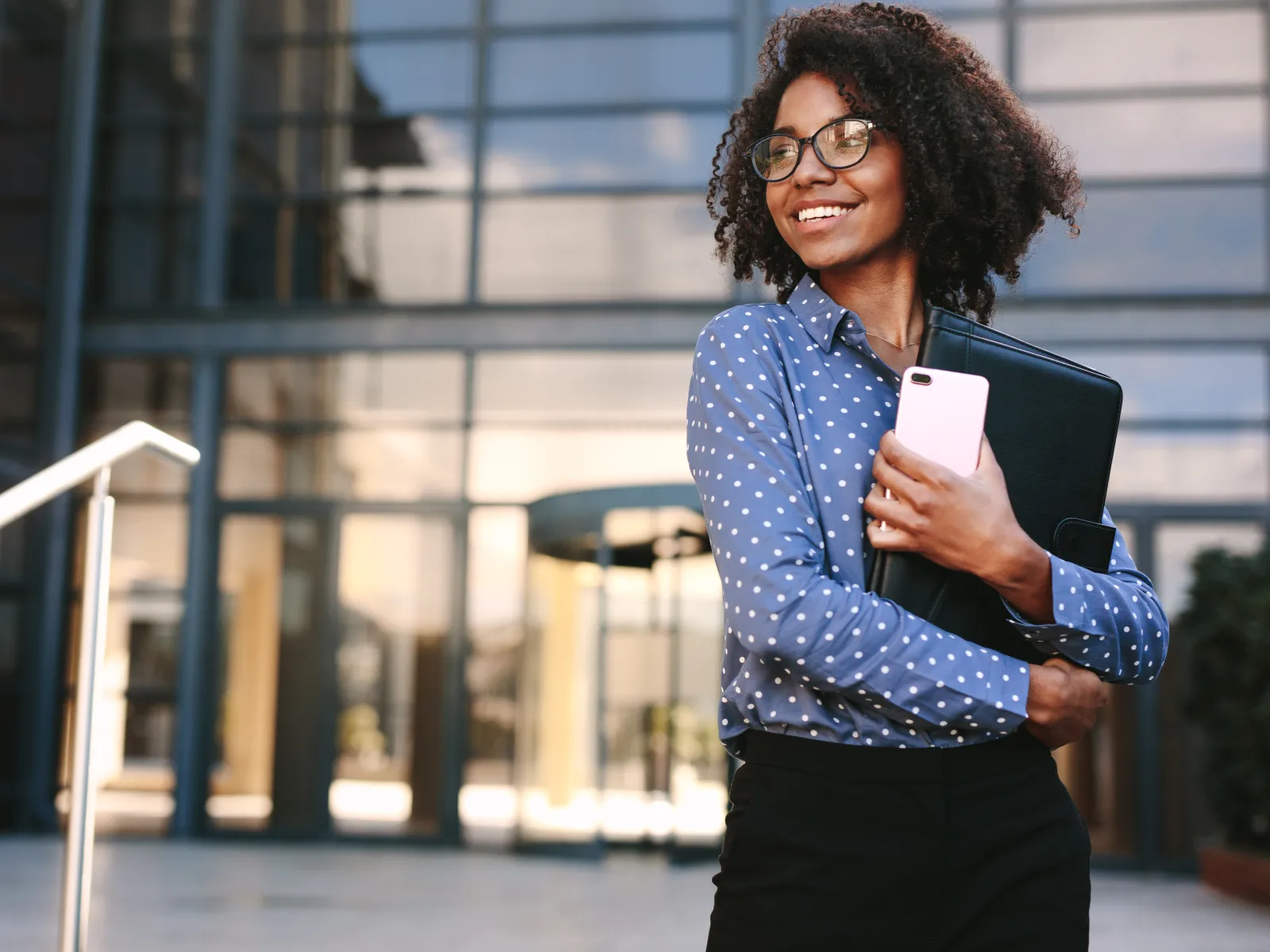 Smiling young woman holding laptop and phone outside modern office building with glass facade.