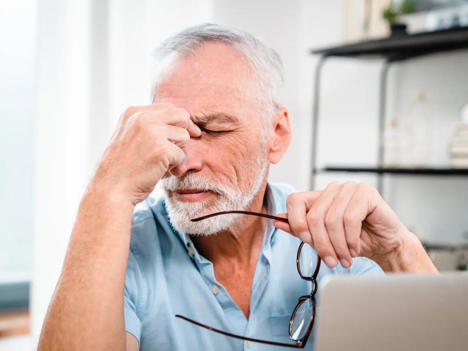 Senior man rubbing forehead and holding glasses, showing signs of stress or headache at a desk with laptop.