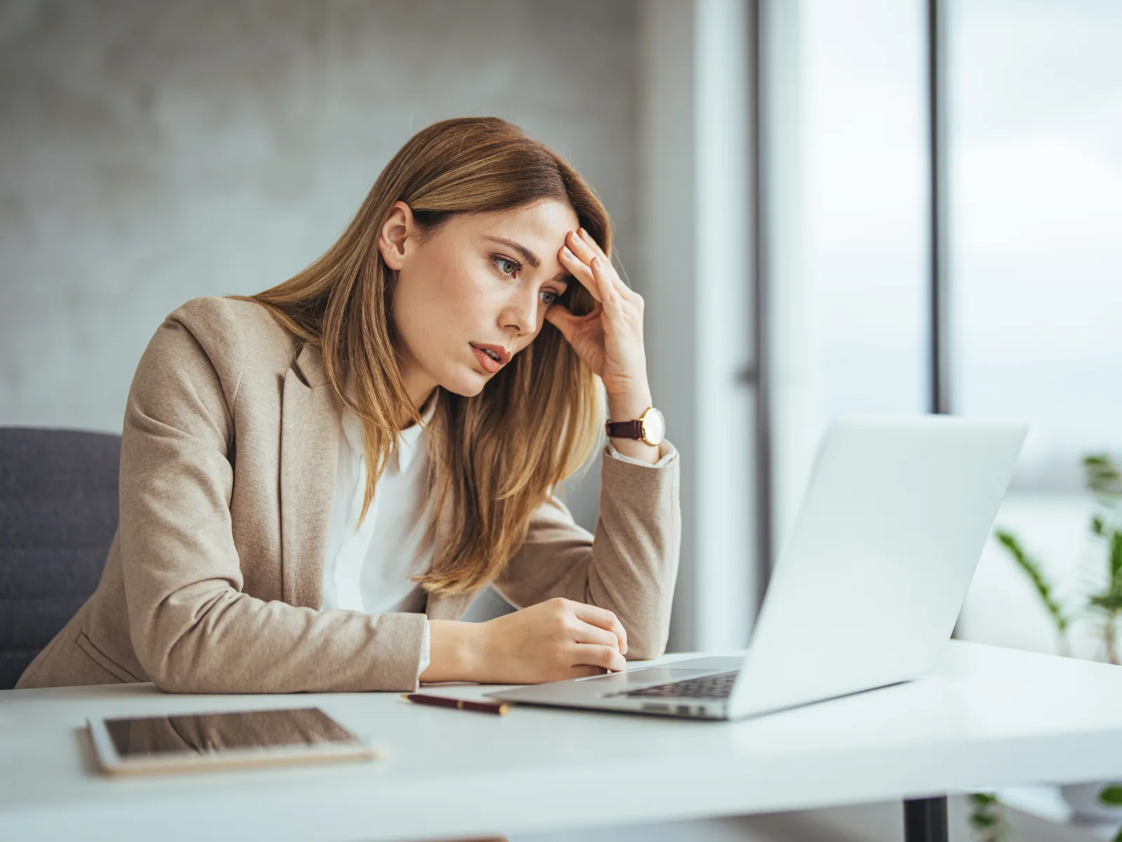 Stressed businesswoman in beige blazer working on laptop at bright office desk with tablet nearby