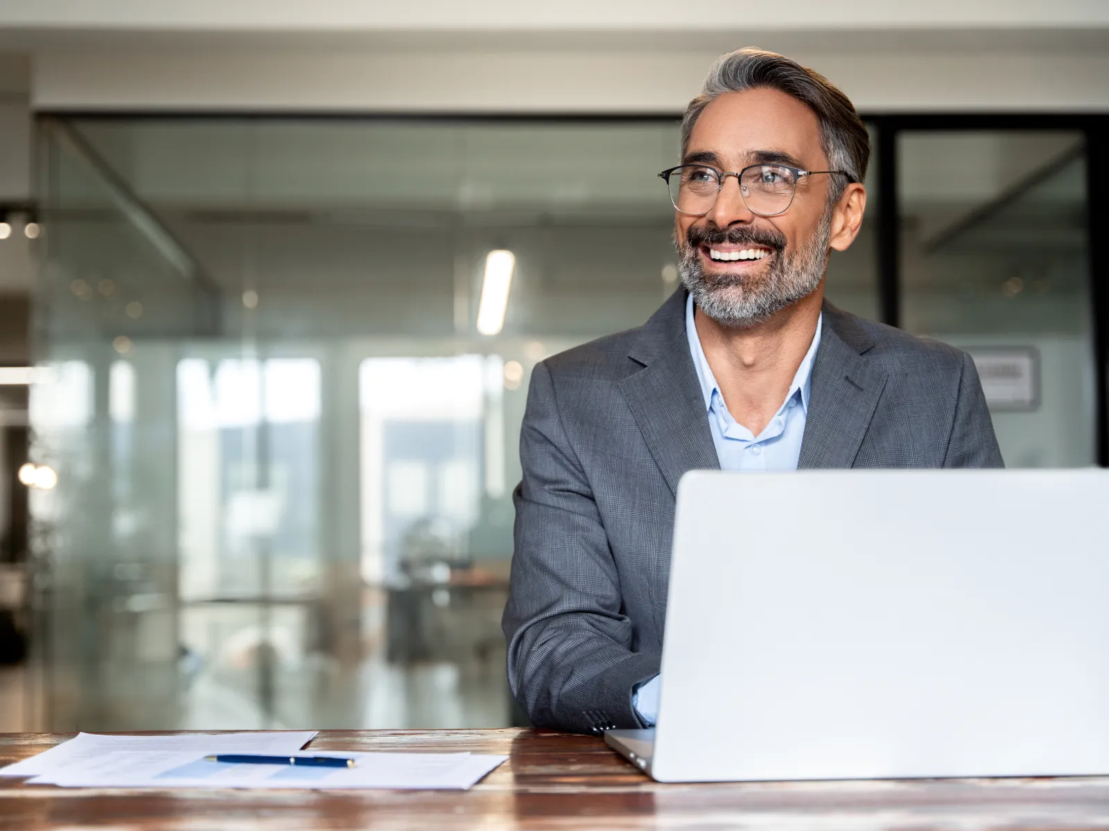 Smiling middle-aged man in a suit working on laptop at office desk with documents and modern glass background