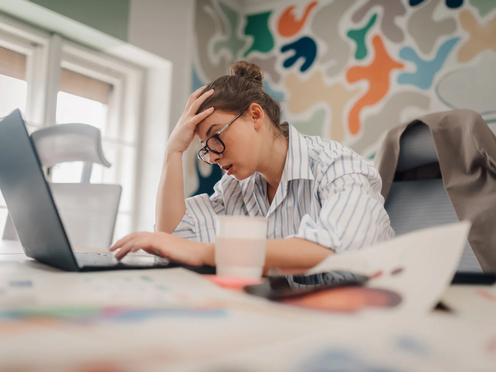 Stressed woman sitting at desk with laptop, holding her head in frustration in a colorful modern office.