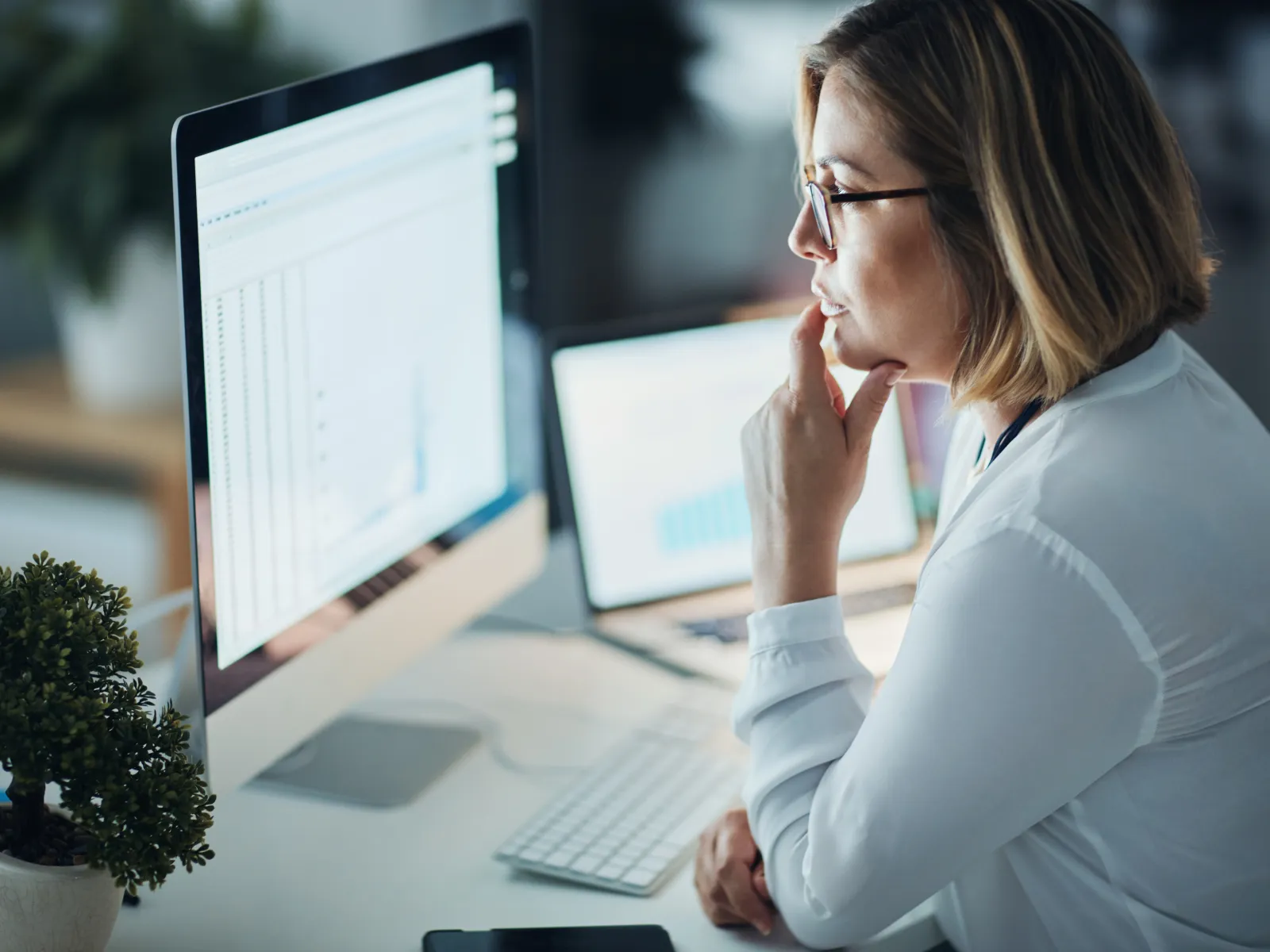 Businesswoman analyzing charts and data on computer screens in a modern office workspace.