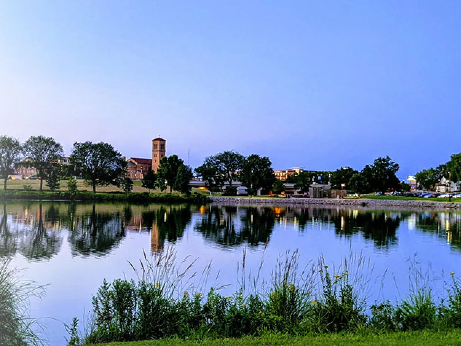 Calm lake reflecting trees and buildings under a clear blue sky with green grass and a paved path in the foreground