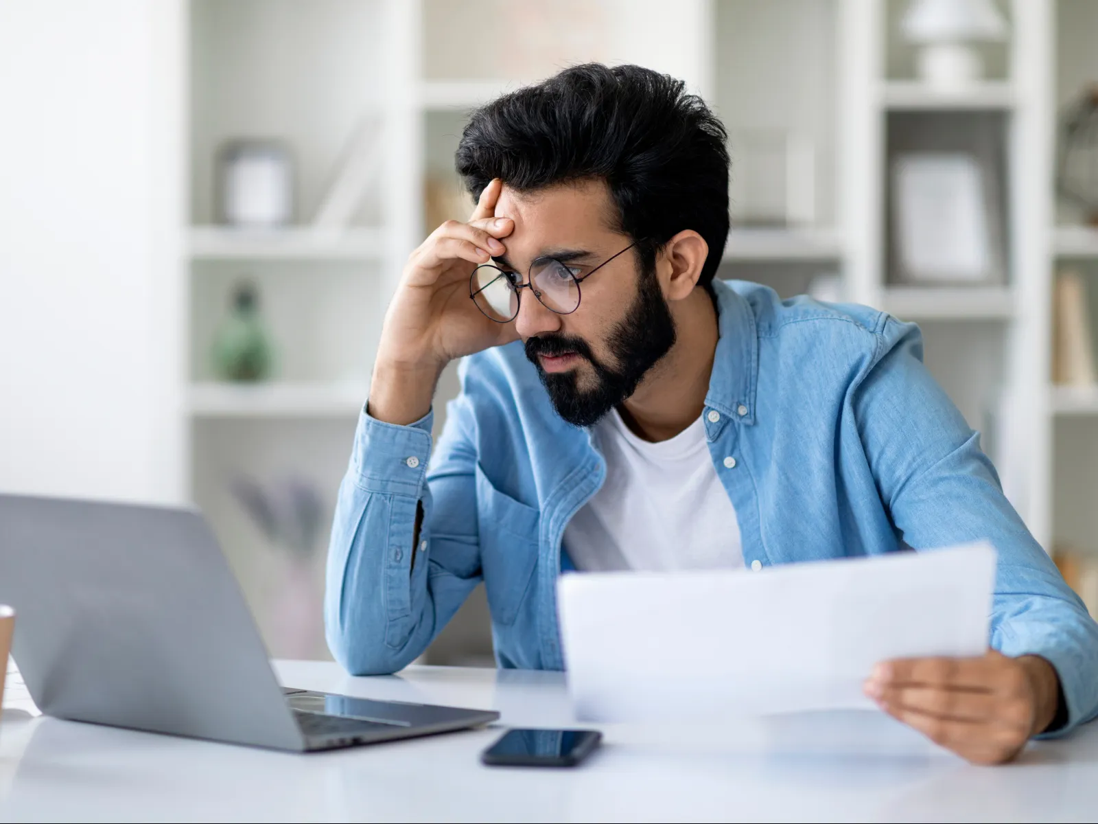 Man with glasses reading documents and working on laptop at home office desk looking concerned.