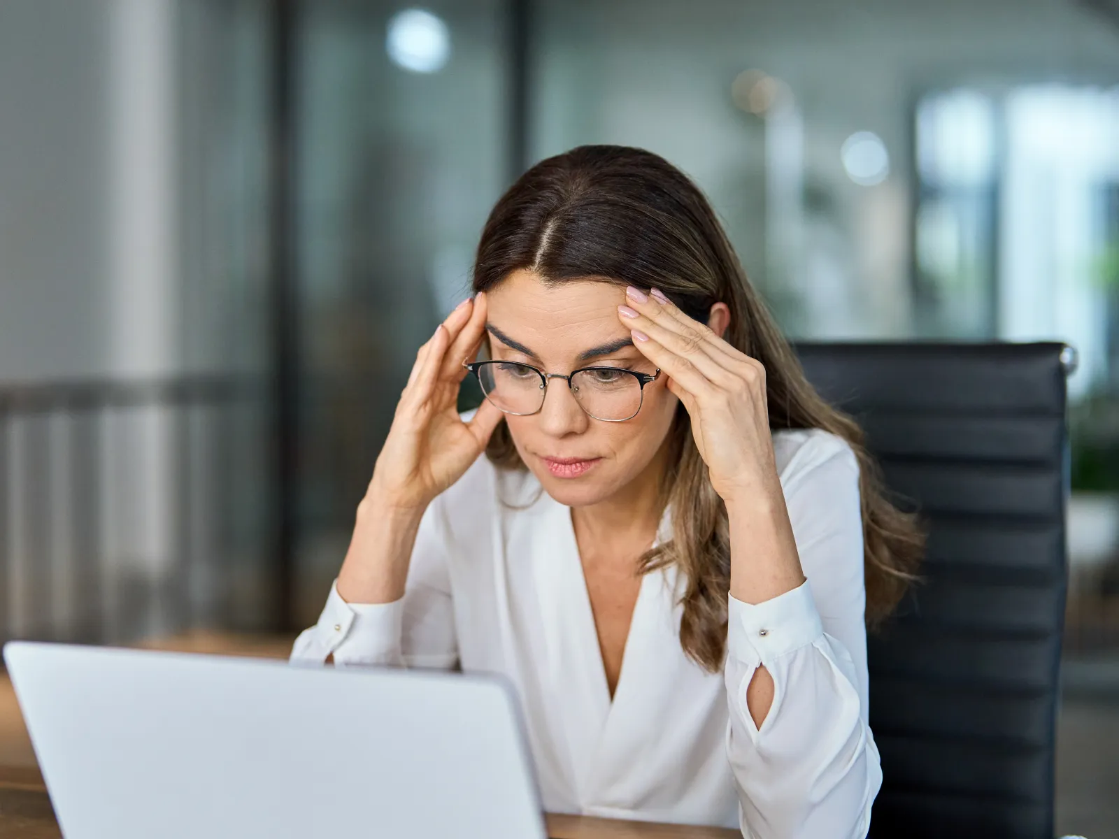 Stressed businesswoman in glasses concentrating intensely on a laptop in a modern office setting.