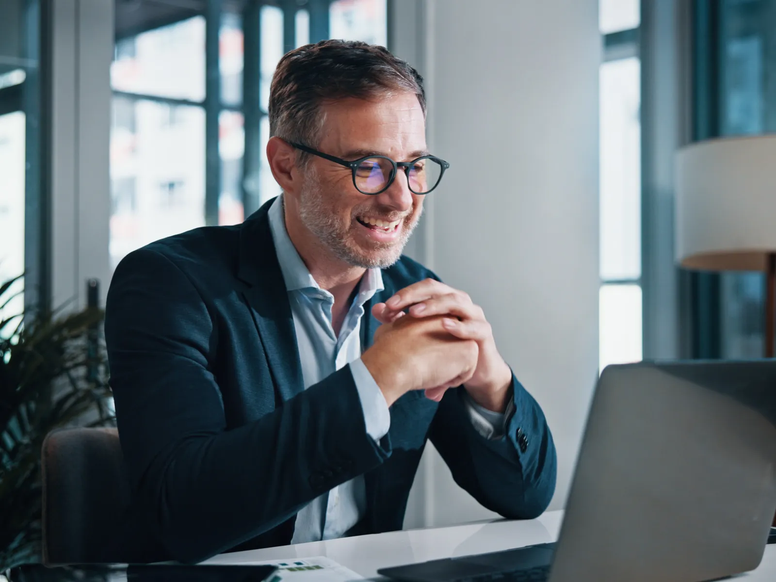 Smiling businessman wearing glasses sitting at desk using laptop in modern office with natural light.