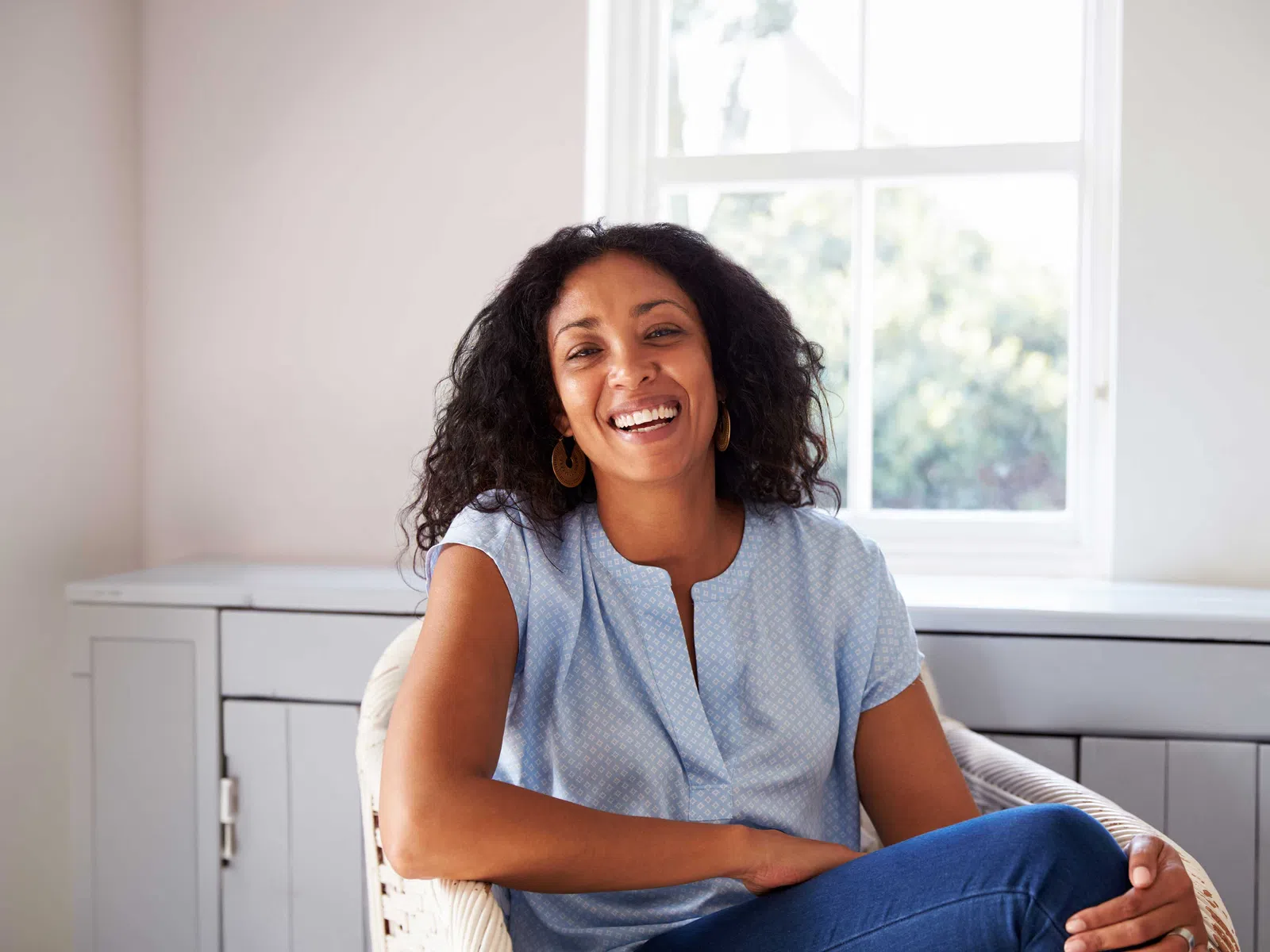 Smiling woman with curly hair wearing a light blue blouse and jeans sitting in a bright room by a window.