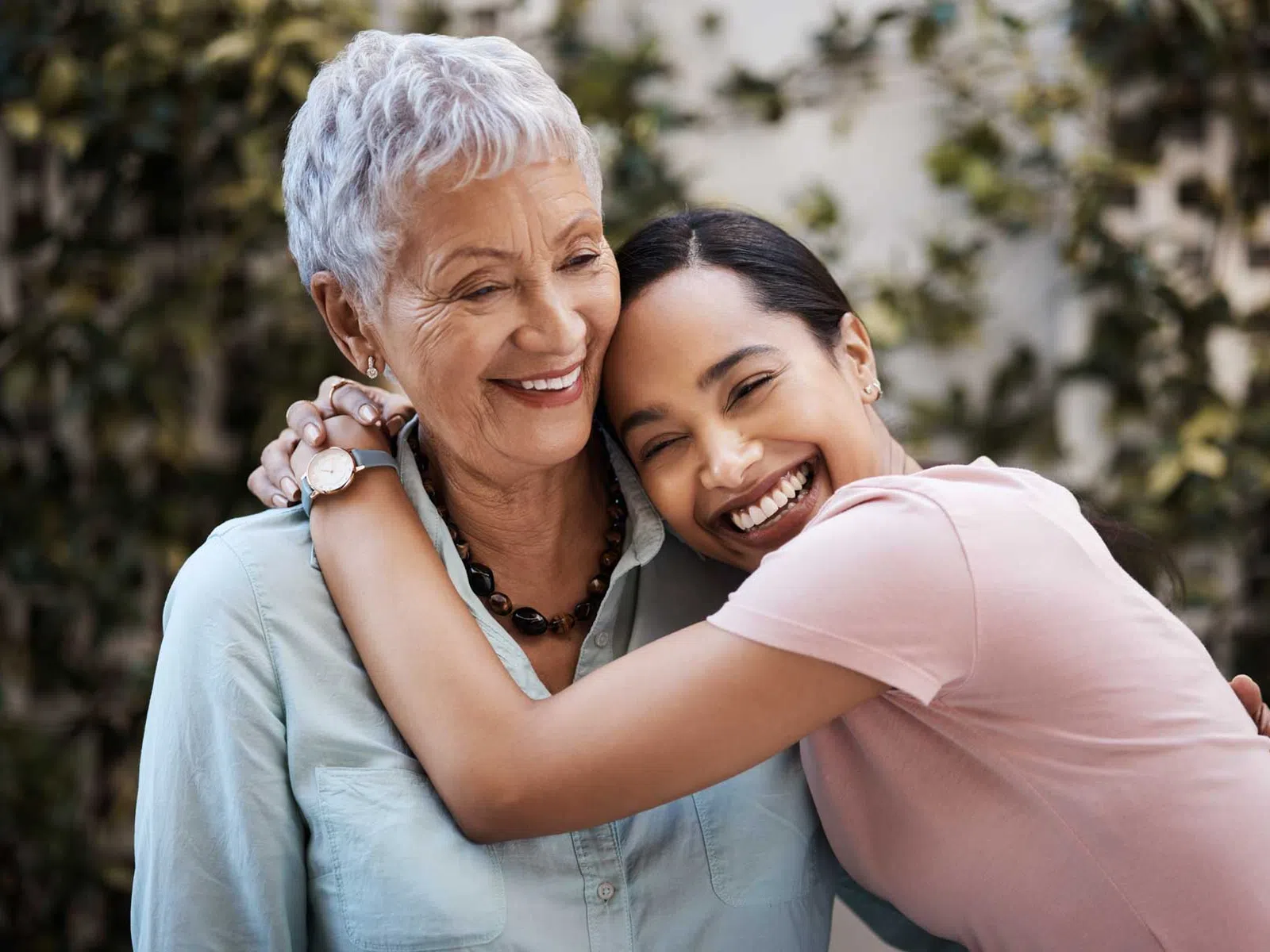 Smiling elderly woman and young woman sharing a warm embrace outdoors with greenery in the background.