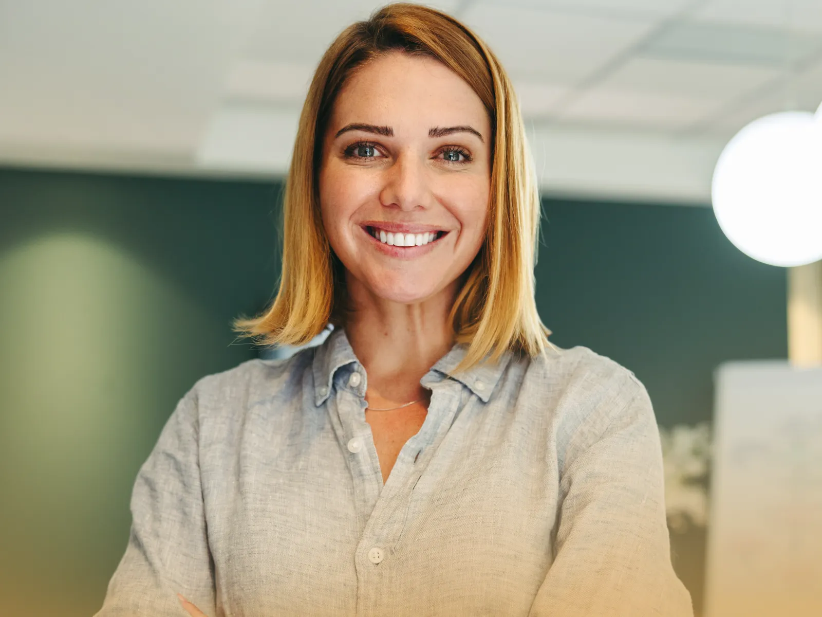 Confident young woman with arms crossed smiling in a modern office with coworkers in background.