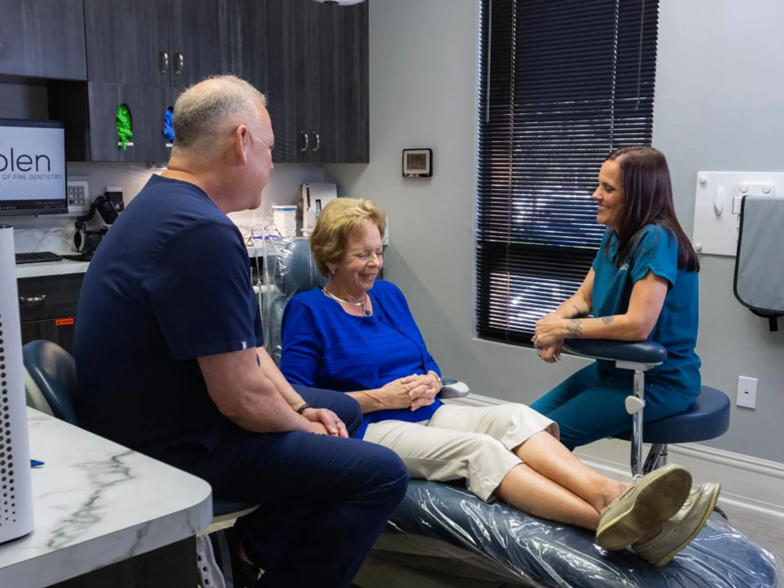 Elderly woman sitting on dental chair smiling with two dental professionals talking in a modern clinic