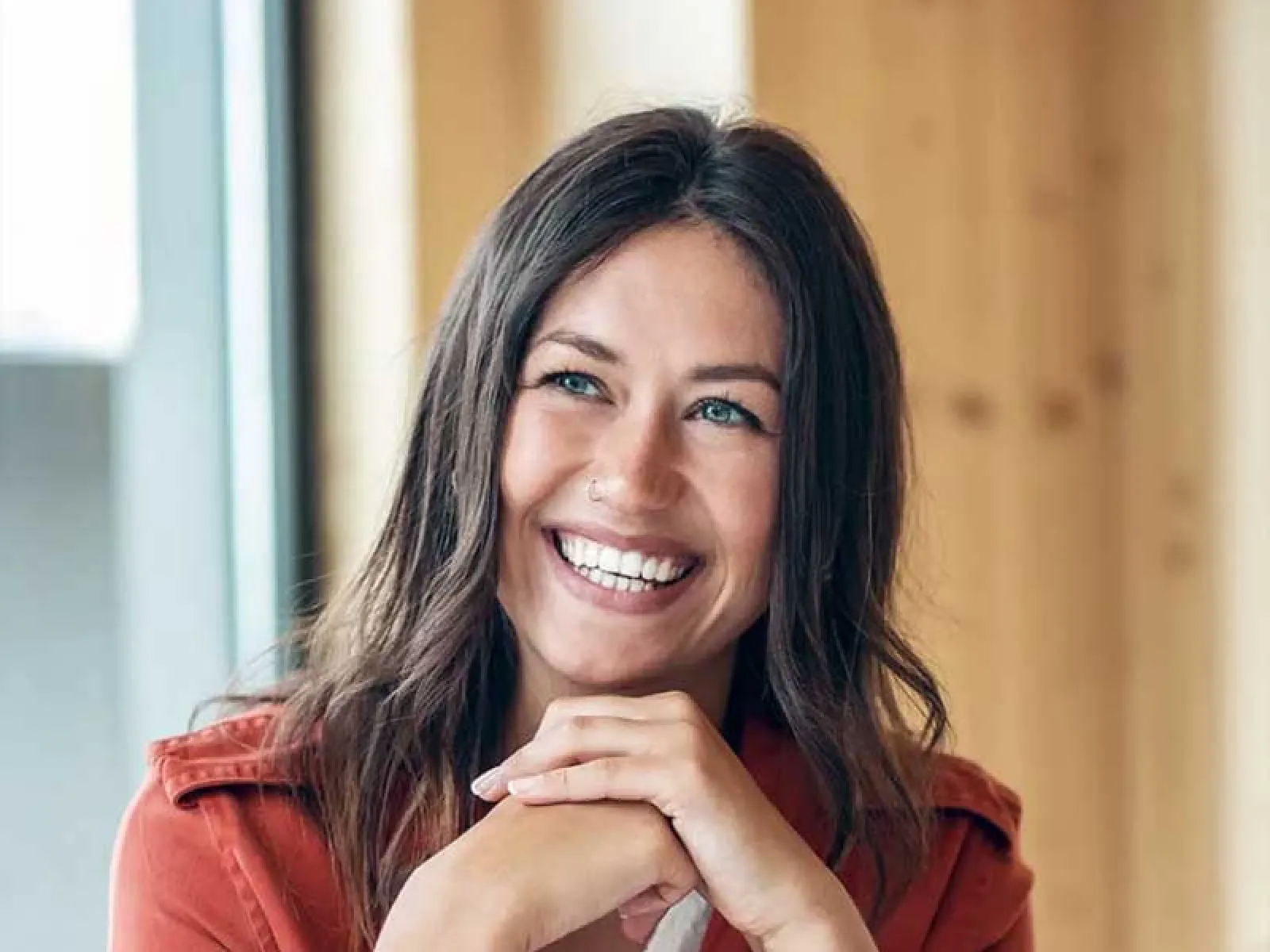 Smiling woman with long brown hair and blue eyes wearing a rust jacket sitting indoors with laptop in front.