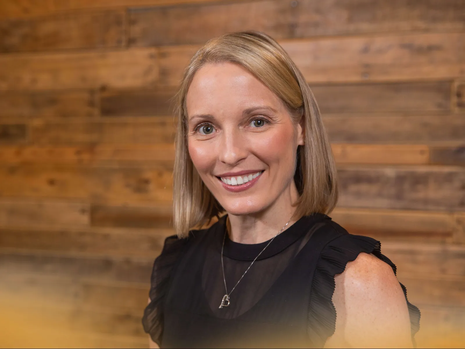 Smiling woman with shoulder-length blonde hair wearing a black top and heart necklace against a wooden background
