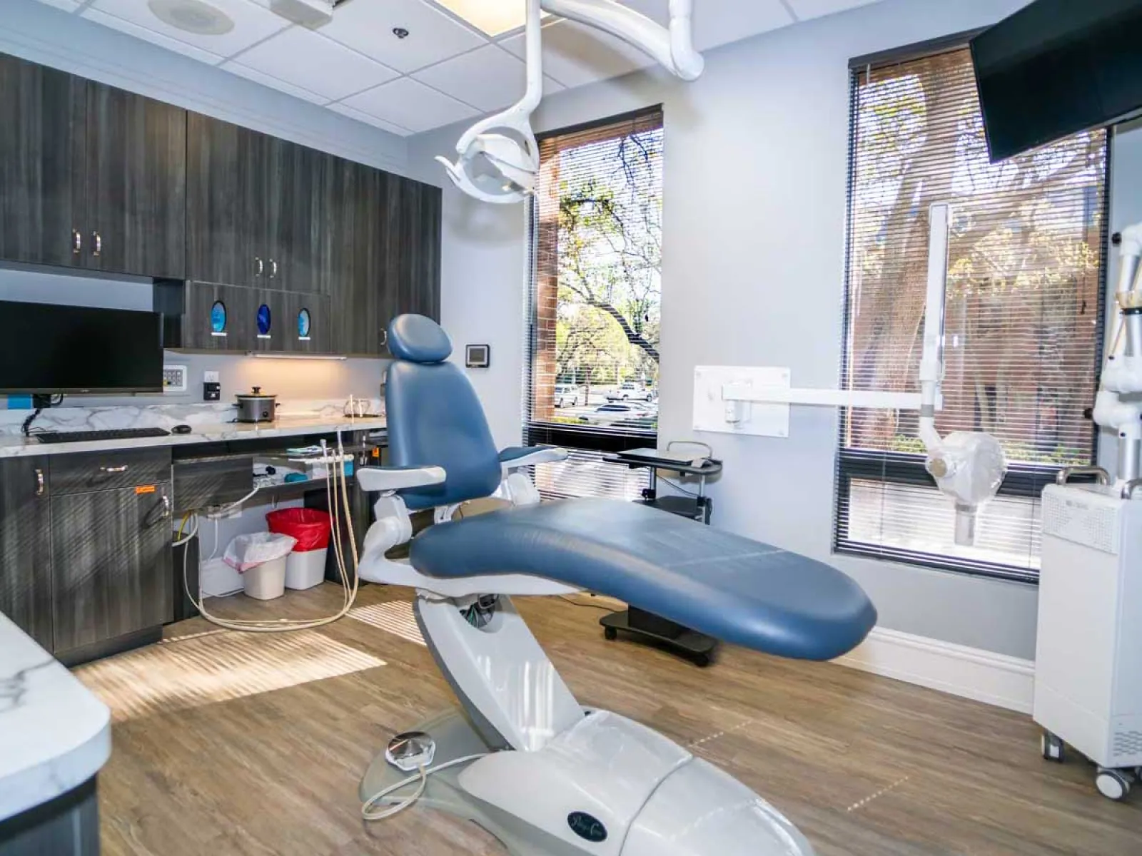 Modern dental clinic room with reclining dental chair, cabinetry, computer monitor, and large window with blinds.