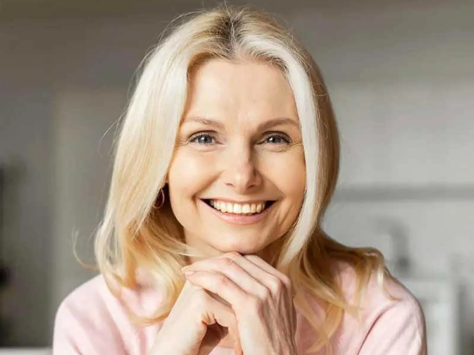 Smiling middle-aged woman with blonde hair wearing a pink sweater, resting her chin on her hands indoors.