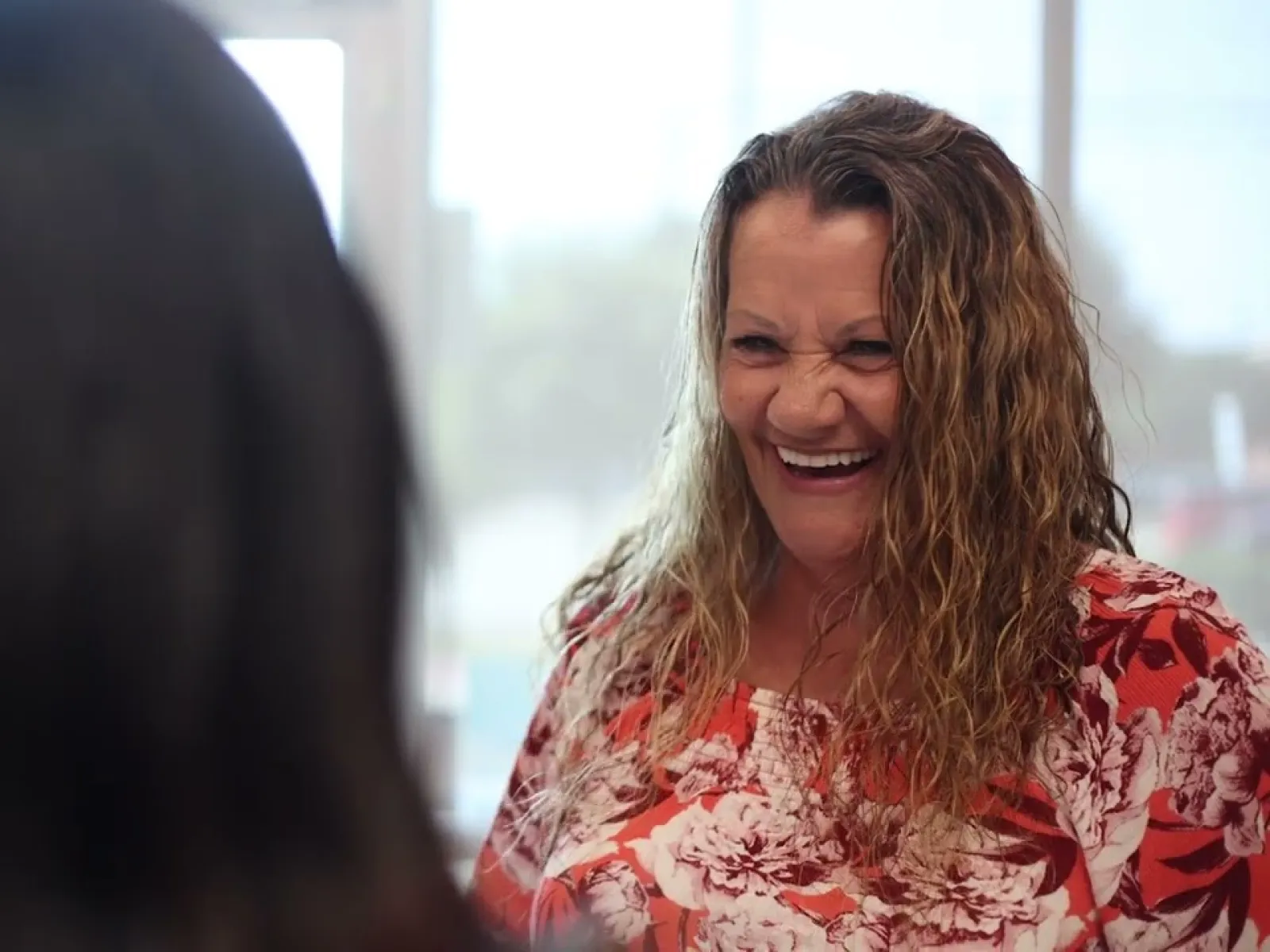 Smiling woman with curly hair wearing a red floral blouse engaged in conversation with another person.