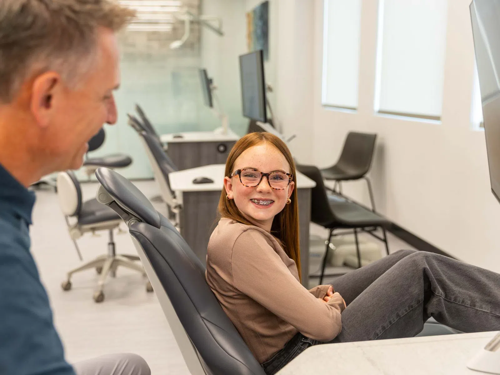 Smiling girl with braces sitting in dental chair talking to dentist in bright orthodontic office.