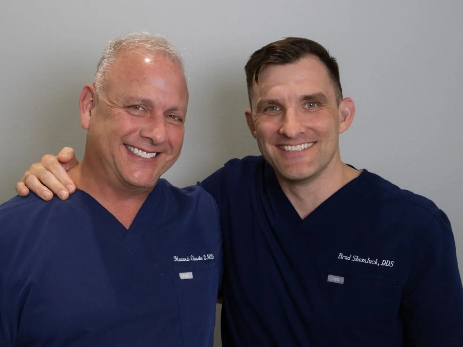 Two male dental professionals in navy scrubs smiling and posing together against a plain background.
