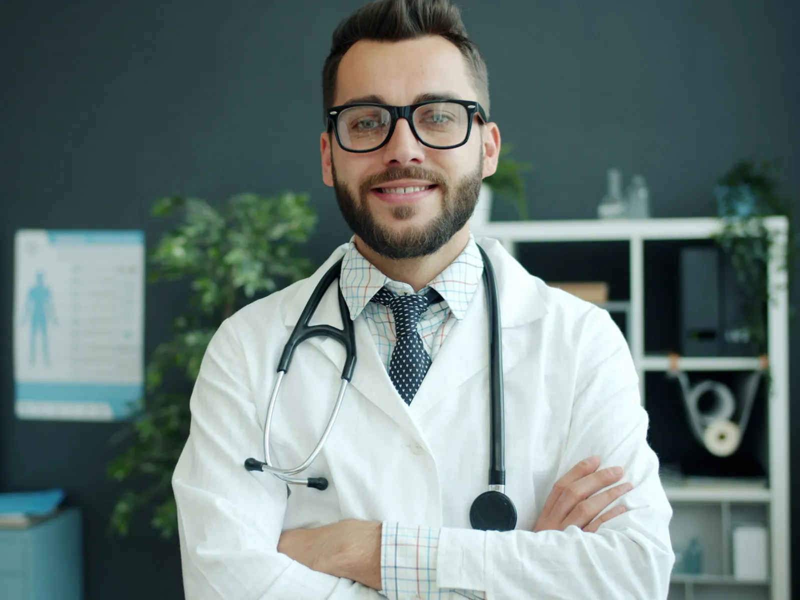 Smiling male doctor wearing white coat and stethoscope standing with arms crossed in medical office.