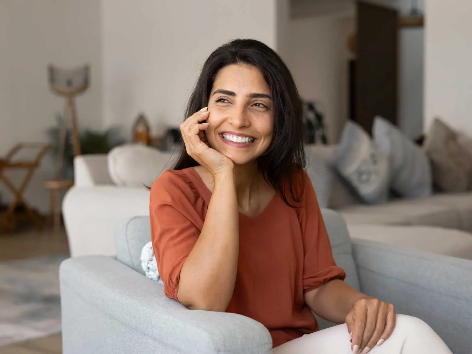 Smiling woman with dark hair wearing an orange shirt sitting on a gray couch in a cozy living room.