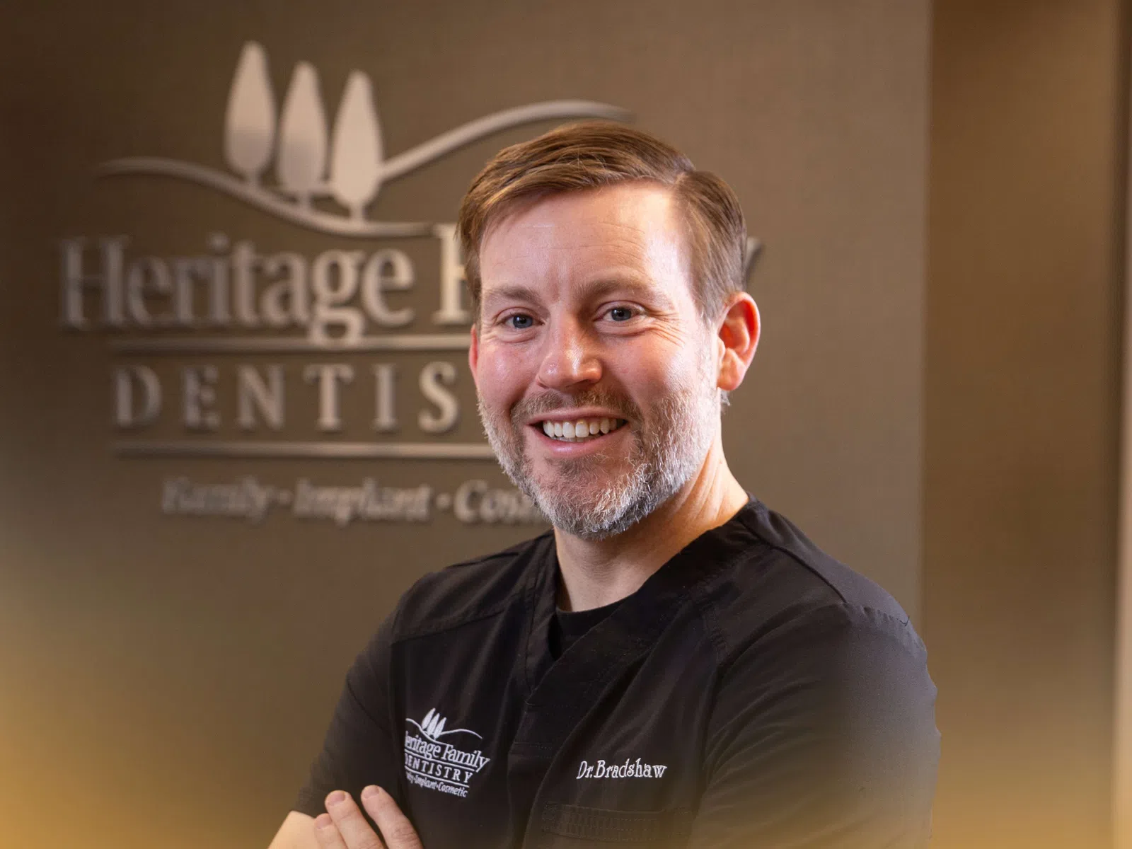 Smiling male dentist Dr. Bradshaw in black scrubs posing in front of Heritage Family Dentistry sign.