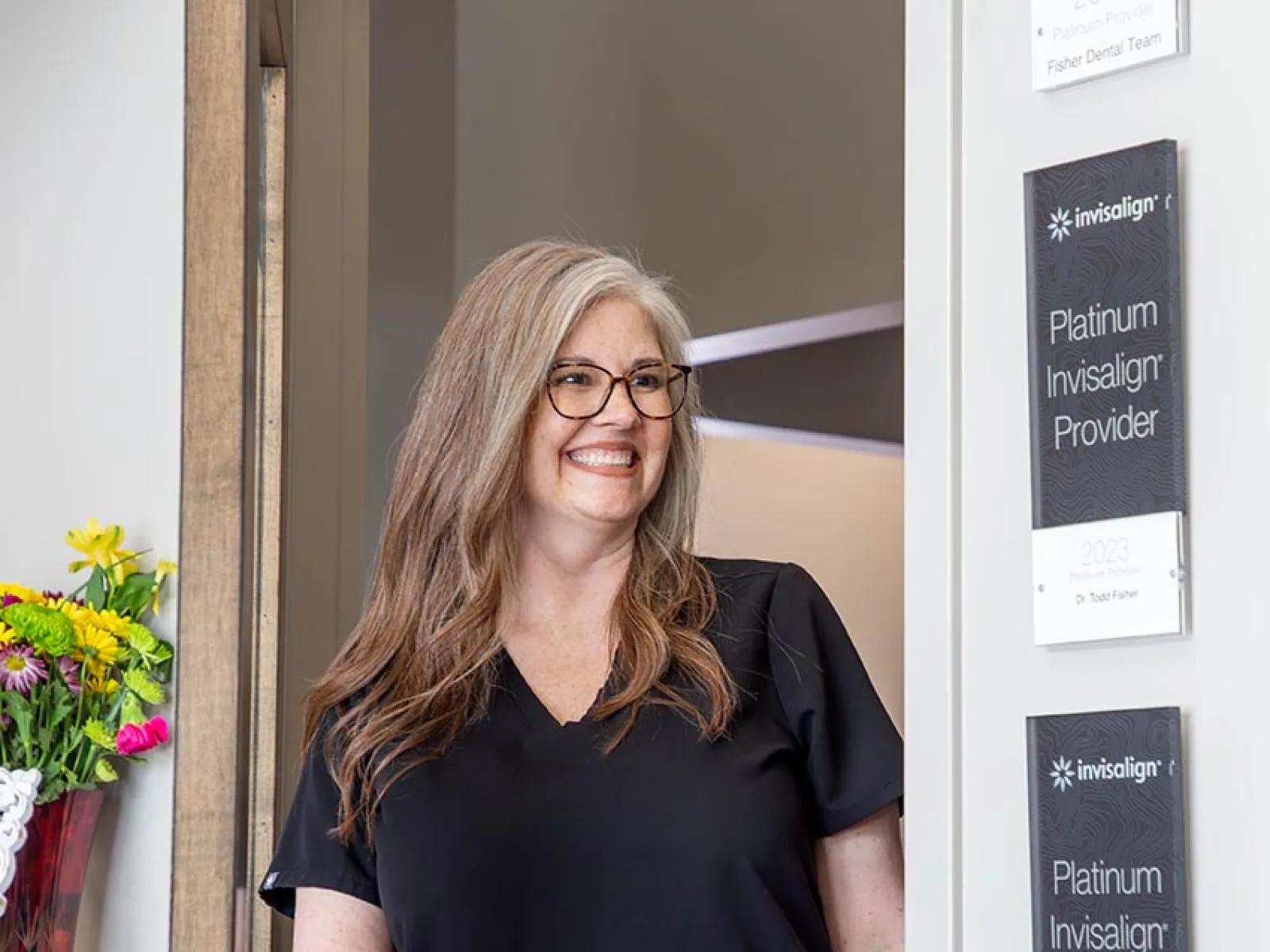 Smiling woman with glasses in black scrubs opening door in dental office with Invisalign provider plaques.