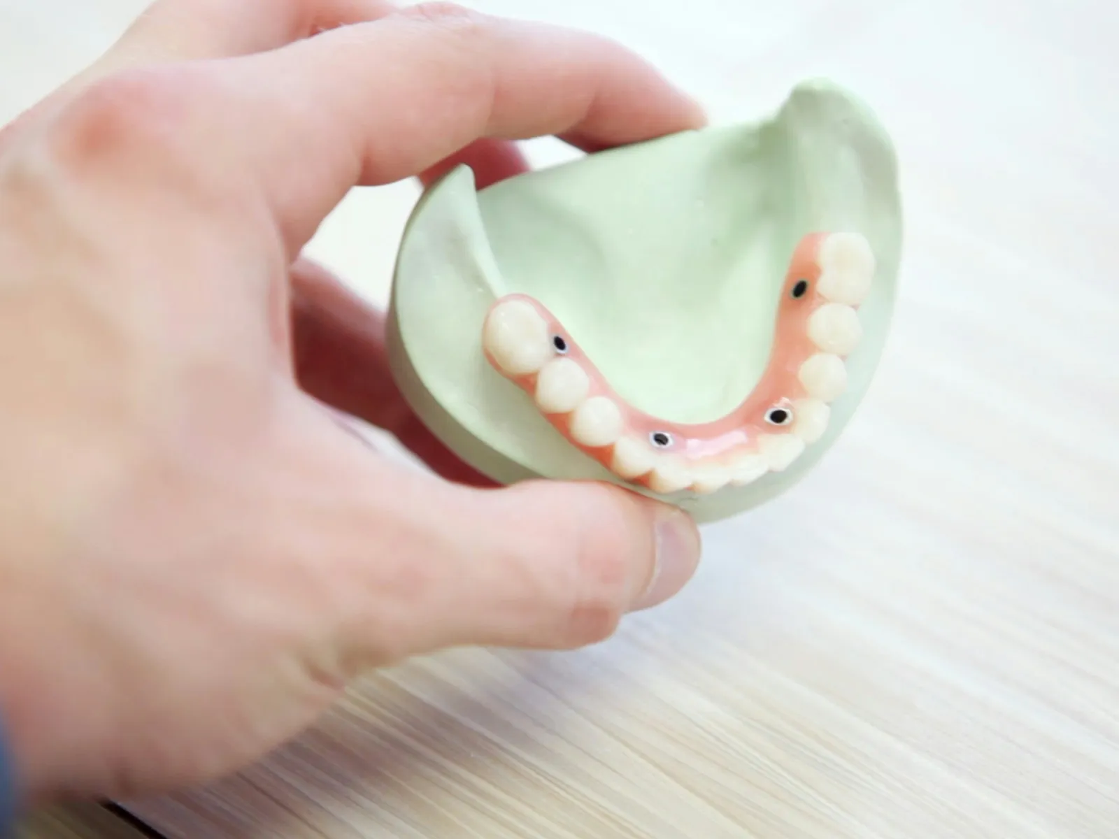 Hand holding dental model showing artificial teeth and implant holes on light wooden surface.