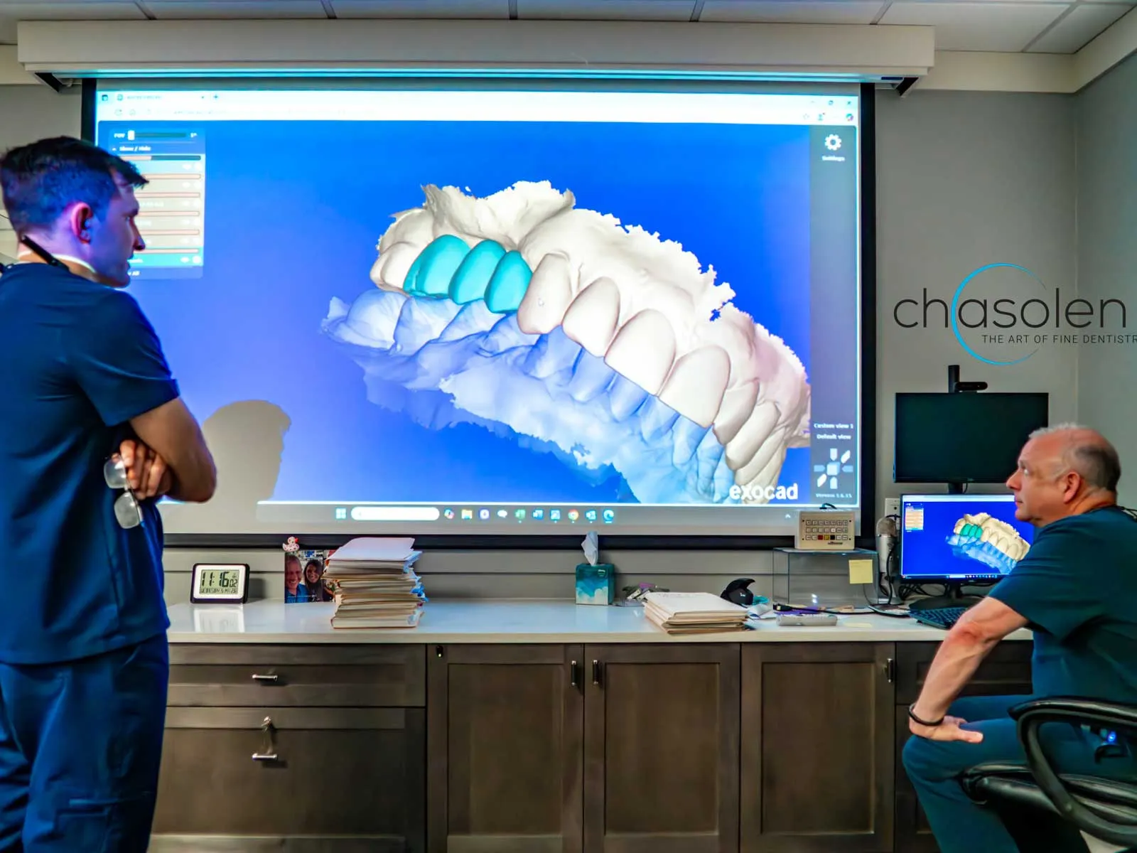 Two dental professionals examining a 3D digital model of teeth on a large screen and computer monitor in an office.
