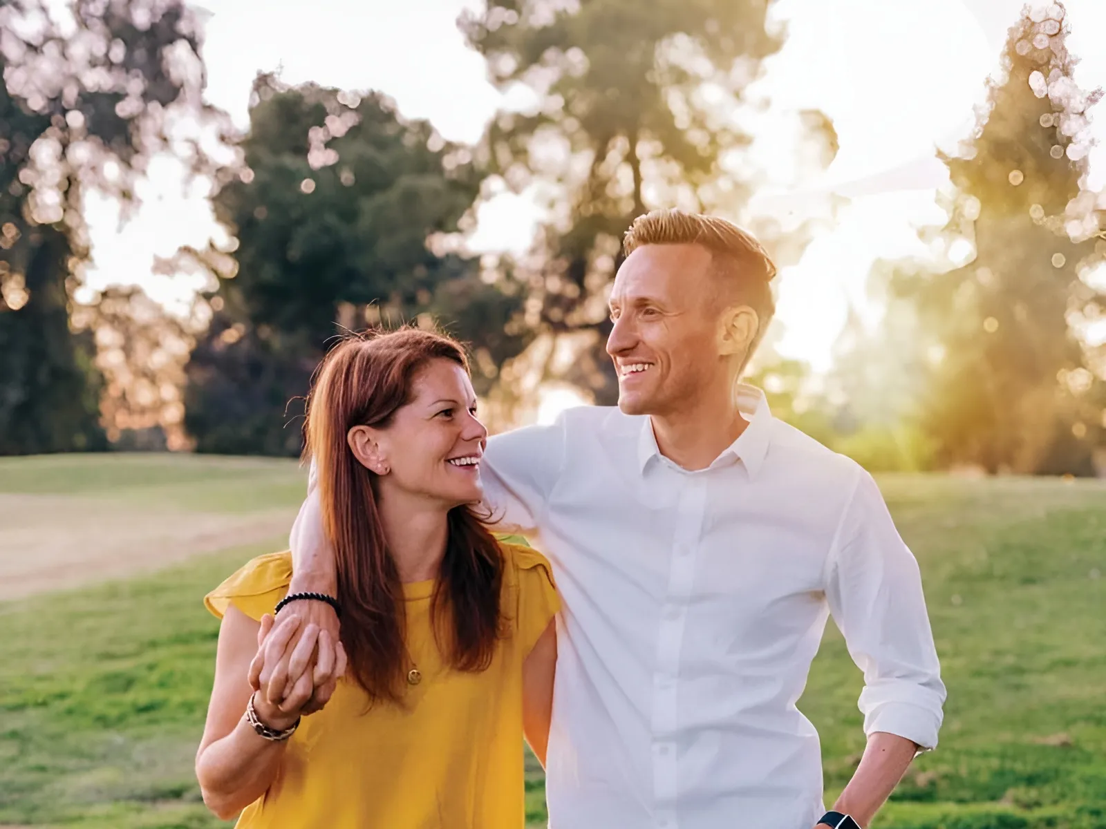 Smiling couple walking outdoors on grass at sunset holding hands and looking at each other happily.